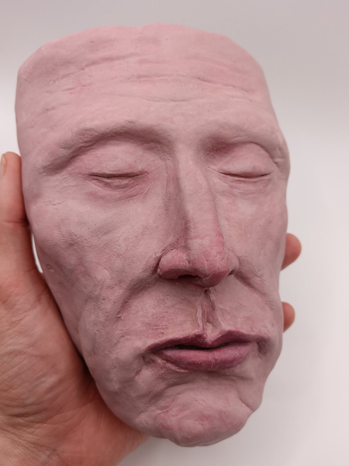 Close-up of a hand holding a pink ceramic face sculpture against a white background