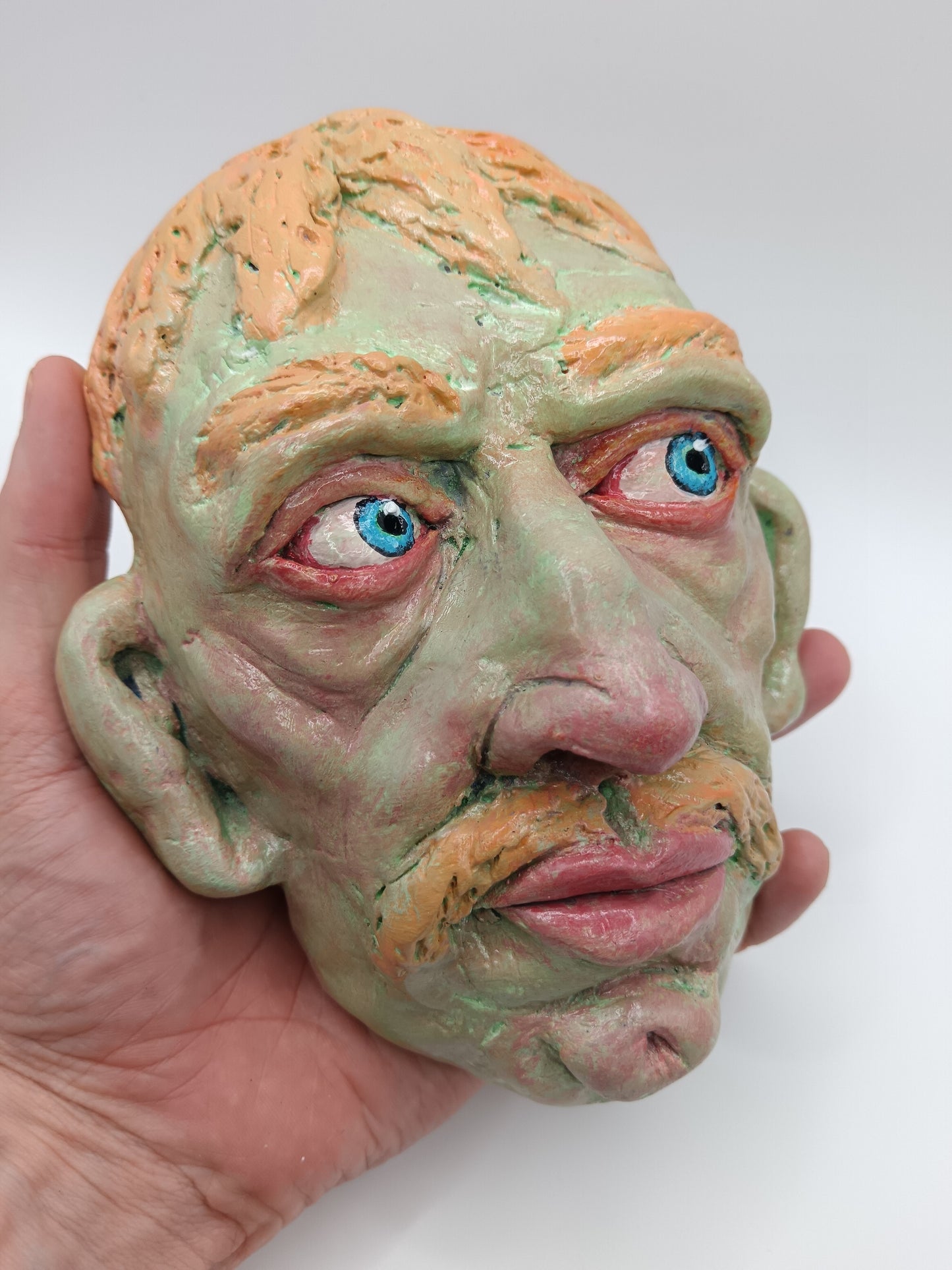 Hand holding a clay sculpture of a face with blue eyes on a white background