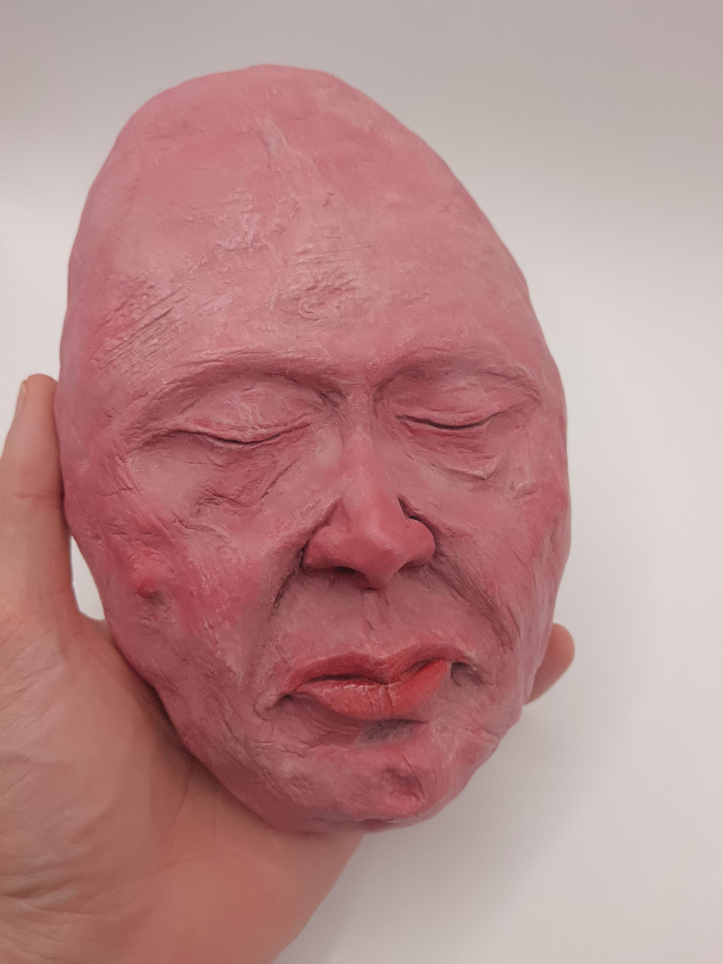 Close-up of a hand holding a pink clay sculpture of a face against a white background