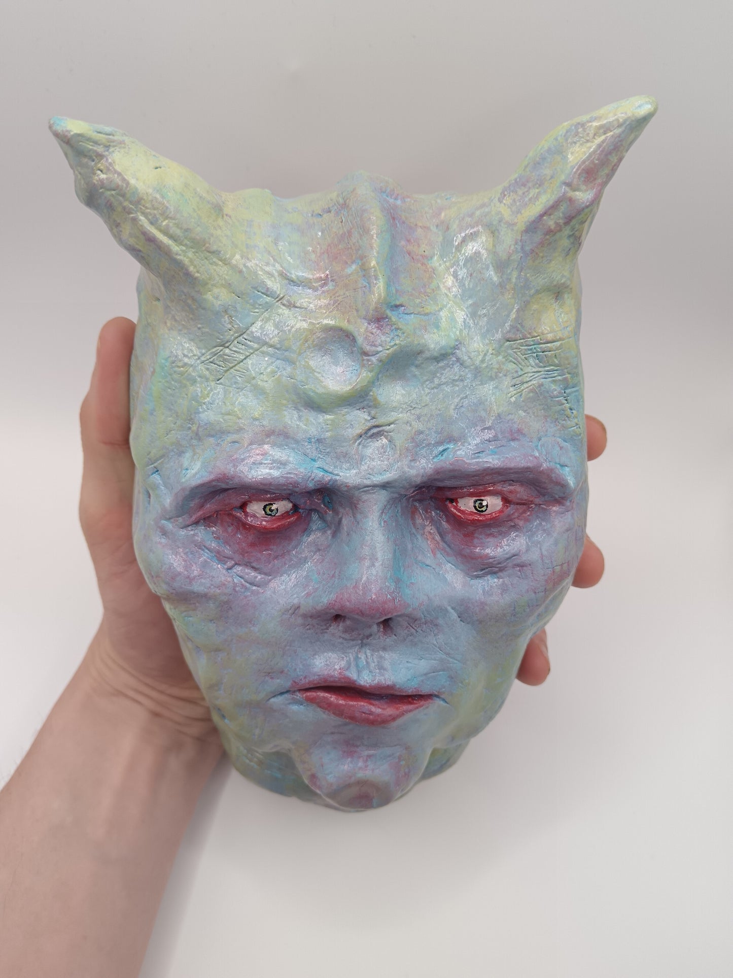 Holographic devil mask with red eyes held by a hand against a white background