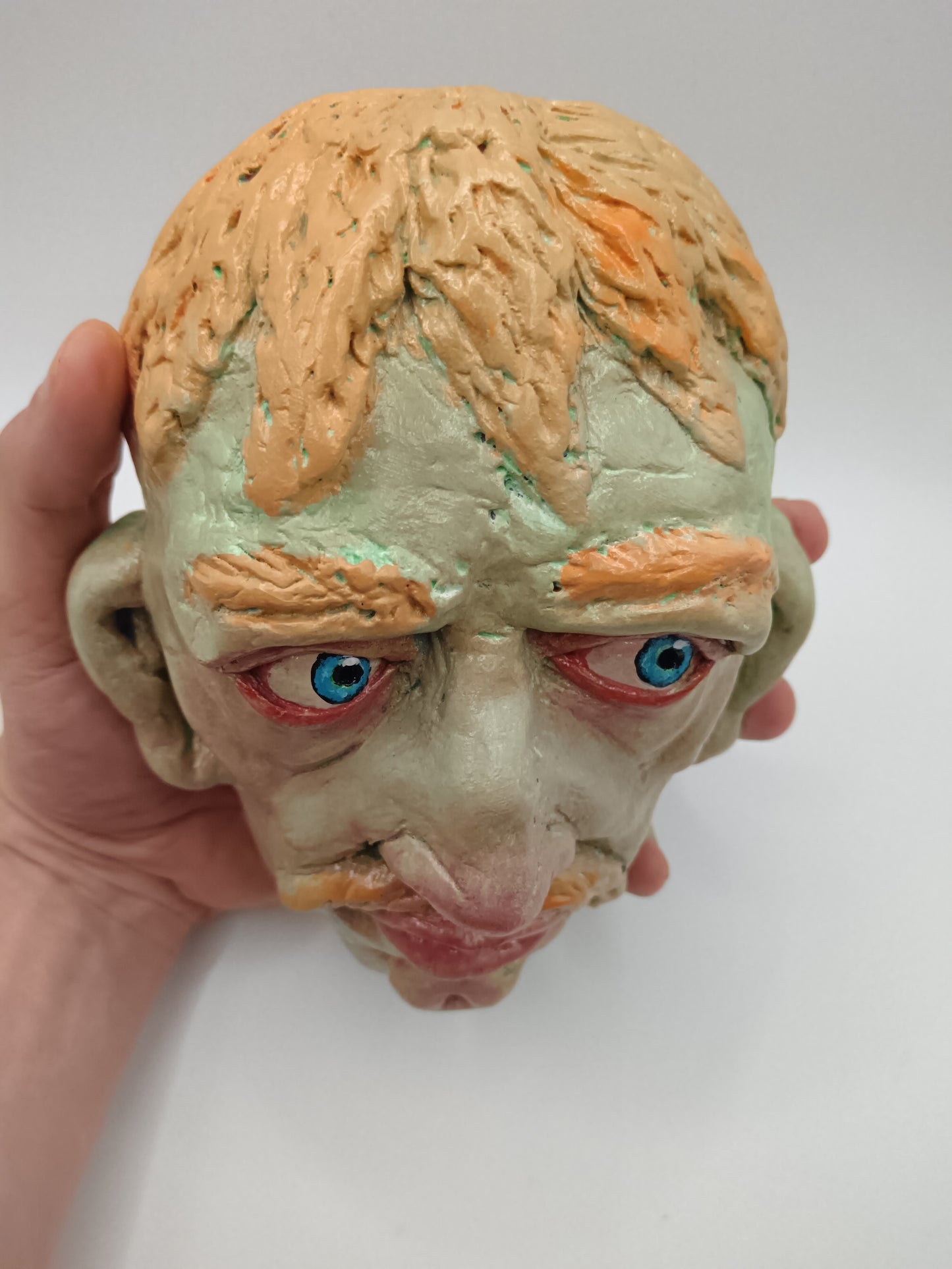 Hand holding a clay sculpture of a face with blue eyes and orange hair against a white background