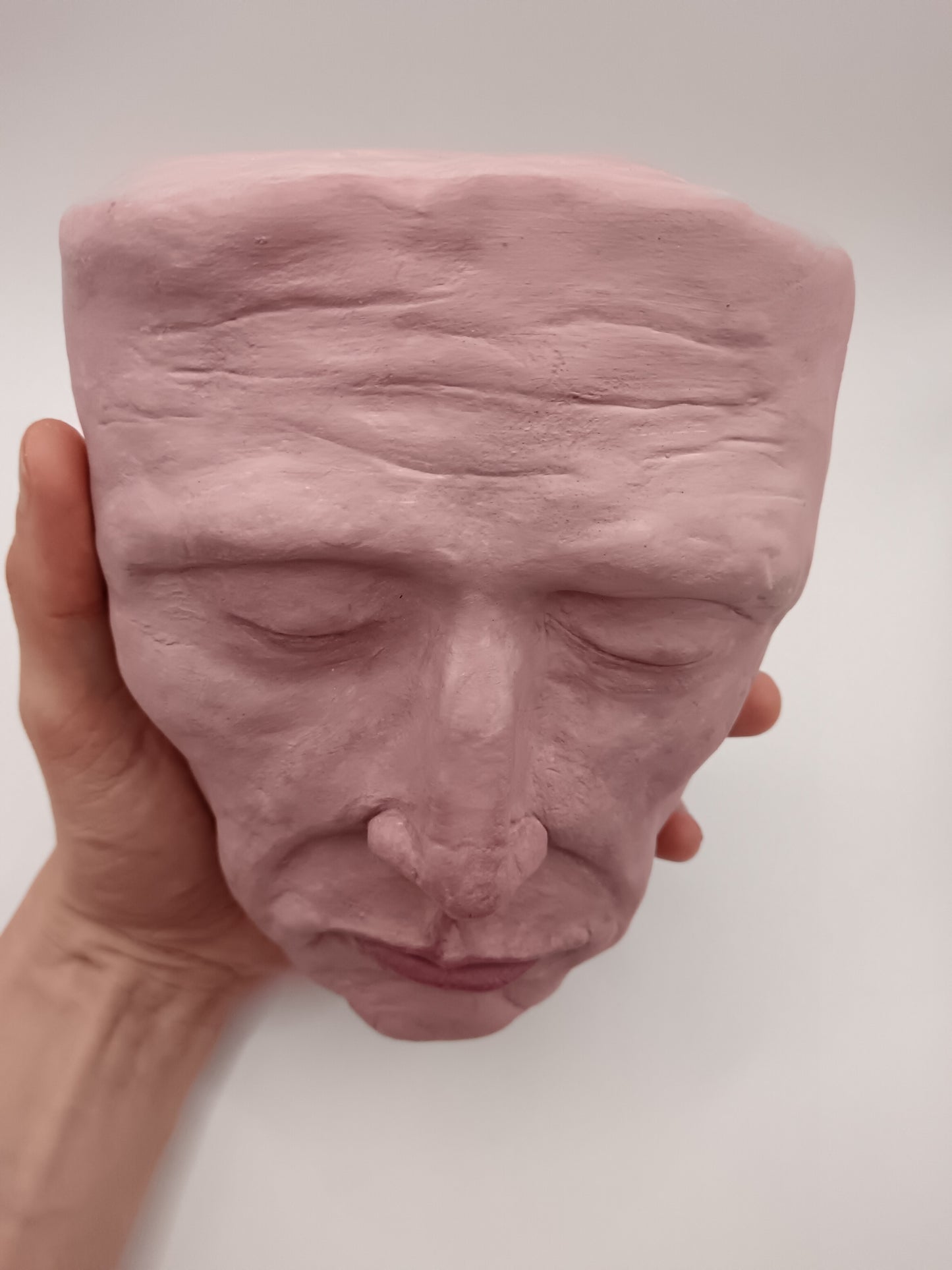 Pink ceramic face sculpture held by a hand against a white background top view