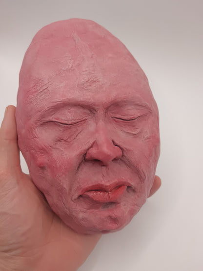 Close-up of a hand holding a pink clay sculpture of a face against a white background