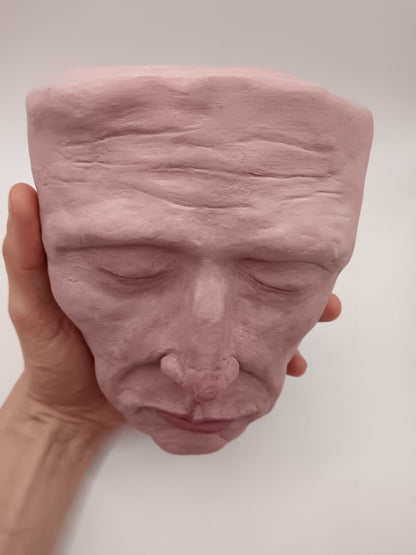 Pink ceramic face sculpture held by a hand against a white background top view