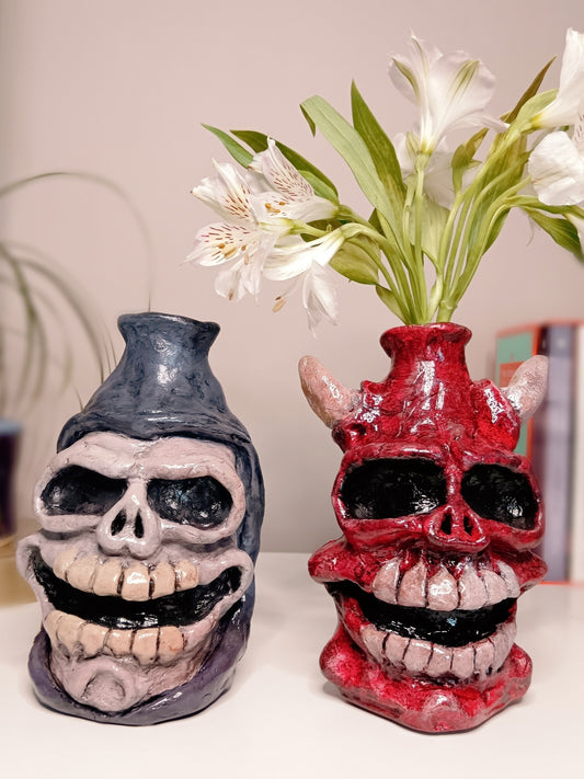 Two skull-shaped vases, one purple and one red, with a vase of white flowers in the background.